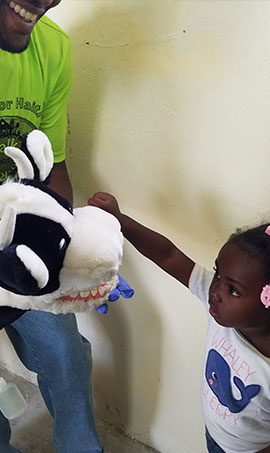 Wearing a blue rubber gloves, a man uses a cow puppet with teeth to let a young child learn how to brush her teeth