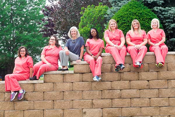 The entire dental team of Georgia Knotek DDS sitting on a brick wall wearing red and Dr. Georgia Knotek wears a black shirt and gray pants in Greenfield, IN