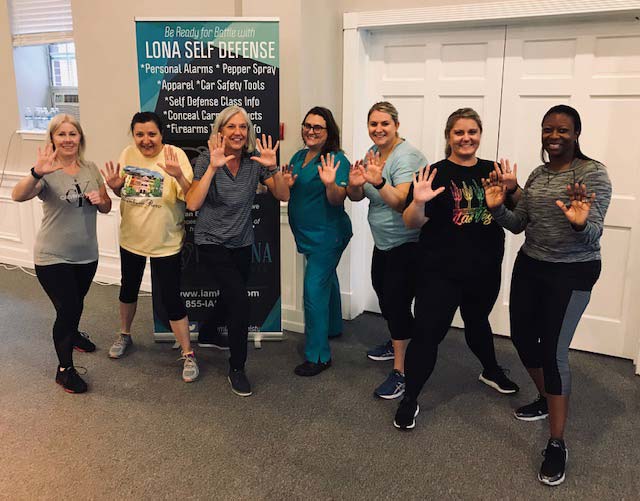 The dental team of Georgia Knotek DDS with their arms and hands raised in front of a Lona Self Defense class banner on a carpeted floor in Greenfield, IN