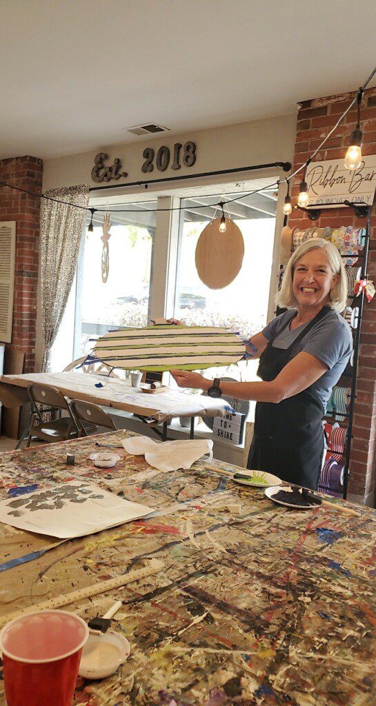Dr. Georgia Knotek DDS standing with her white, blue, and green sign at a local art studio next to a well used paint table in Greenfield, IN