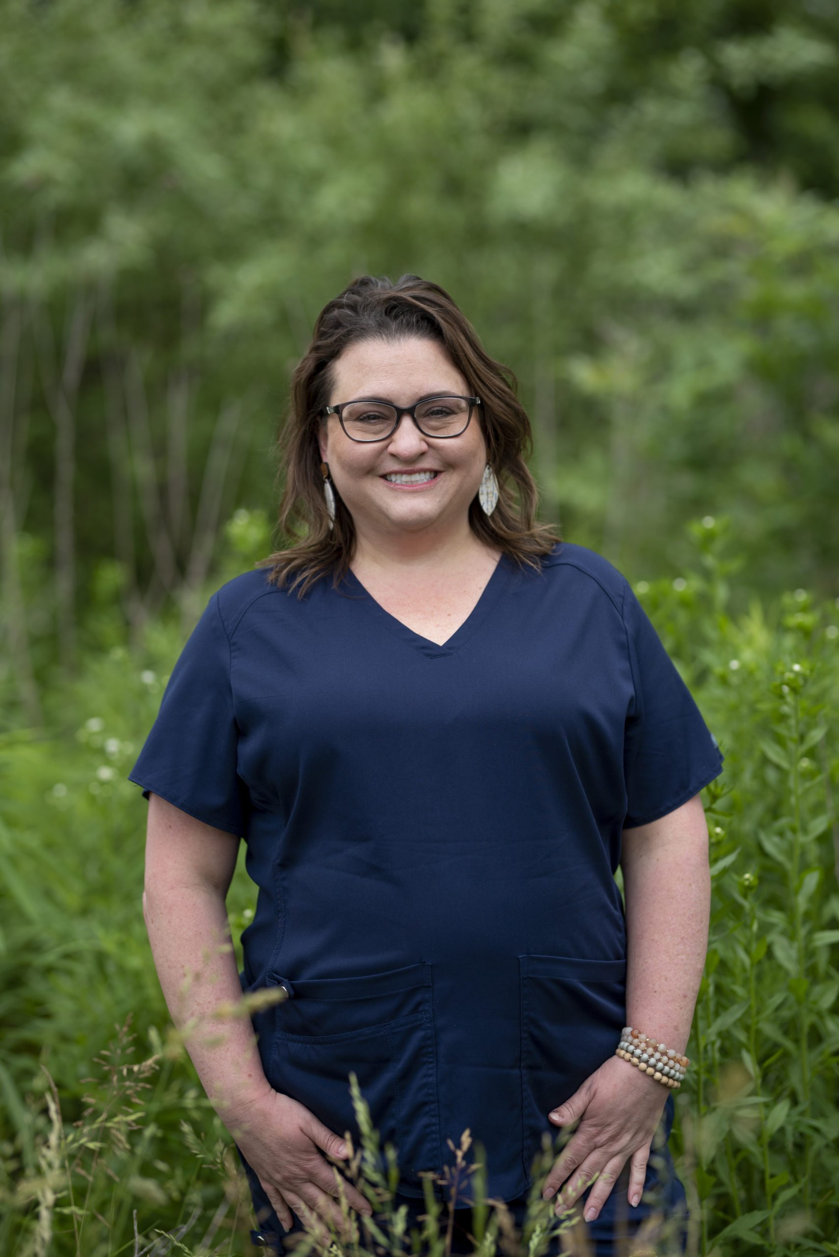 Carrie, dental assistant, wearing a blue shirt and black glasses amid a lush green forest in Greenfield, IN