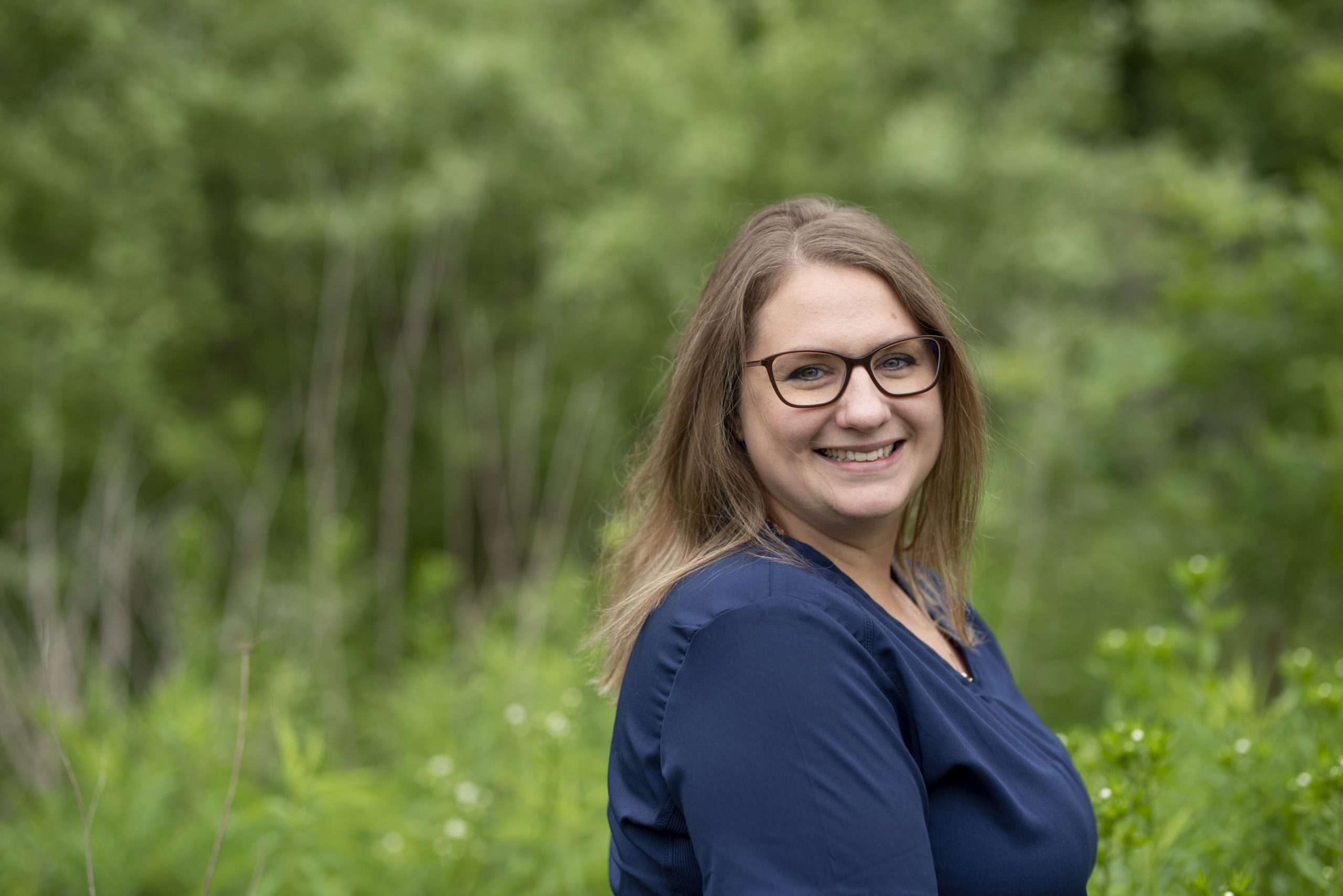 Kathleen wearing a blue shirt and brown glasses in front of a lush green forest in Greenfield, IN